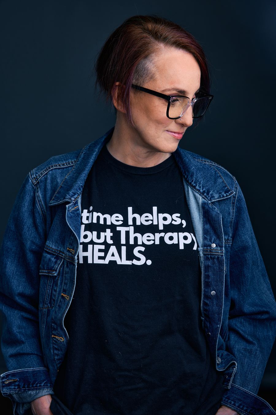 Alex Fliess, LCSW, in a denim jacket and a T-shirt that reads "time helps, but therapy heals."